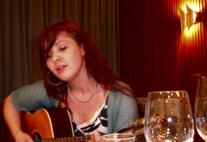 Rebekah Del Rio playing acoustic guitar in a warmly lit room, captured during an intimate performance.