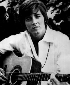 Bobby Sherman strumming an acoustic guitar in a vintage photo, wearing a casual shirt and smiling.