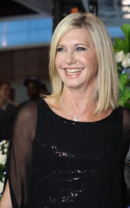 Olivia Newton-John smiling warmly in a black outfit during a photoshoot, exuding classic elegance.